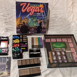 May include: A board game titled "Lords of Vegas" with various game components, including dice, cards, and game boards. The box art features a cityscape with the game's title in a stylized font. The game pieces are in black trays.