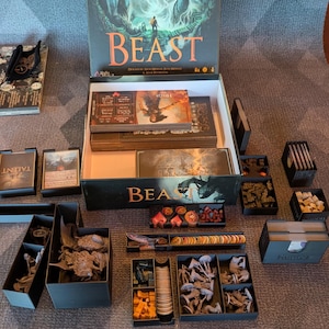 Puede incluir: Juego de mesa "BEAST" con una escena de bosque en la caja. El juego está abierto, revelando cartas, fichas y figuras en miniatura en contenedores de almacenamiento negros. El juego está sobre una mesa.