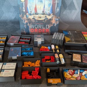 Può includere: Un gioco da tavolo intitolato World Order con una scatola che raffigura una riunione diplomatica. Il set include diversi vassoi in plastica nera riempiti con vari pezzi di gioco colorati, tra cui gettoni rossi, gialli, blu e bianchi, insieme a mazzi di carte e tabelloni.