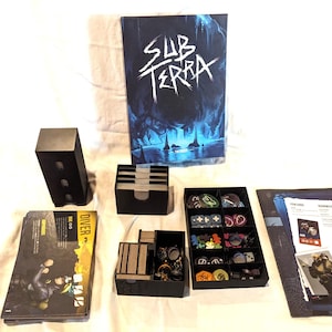 Könnte beinhalten: Ein Brettspiel-Set mit einer Box, die "SUB TERRA" in weißer Schrift auf einem dunkelblauen und schwarzen Hintergrund zeigt. Das Set enthält schwarze Aufbewahrungsboxen, Karten und Spielsteine.