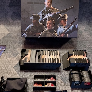 Może przedstawiać: Pudełko gry planszowej Sniper Elite z trzema żołnierzami z karabinami, w zestawie z czarnymi plastikowymi organizerami zawierającymi karty, okrągłe żetony, małe kolorowe kostki, czarny woreczek oraz czarny długopis.