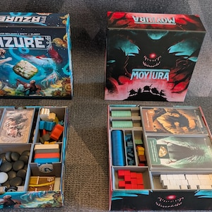 Puede incluir: Dos cajas de juegos de mesa, "Azure" y "Moyura", con organizadores de componentes de juego abiertos. La caja "Azure" presenta una escena acuática azul y dorada. La caja "Moyura" tiene un diseño de monstruo rojo y negro. Los organizadores contienen piezas de juego.