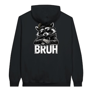Puede incluir: Sudadera con capucha negra con un gráfico de un mapache con gafas de sol y gorra, con la palabra "BRUH" en blanco. El mapache está representado en una pose relajada, dando a la sudadera un ambiente urbano.