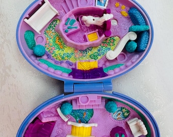Puede incluir: Un set de juego de plástico morado con forma ovalada que se abre en dos niveles. El nivel superior cuenta con una pequeña habitación con cama y un jardín. El nivel inferior contiene un estanque con un puente amarillo, un caballo y muebles.