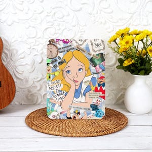 May include: A rectangular decorative item featuring Alice from Alice in Wonderland. The design includes Alice with blonde hair and a blue dress, along with text and illustrations. It rests on a woven mat, with a vase of yellow flowers in the background.