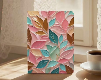 Puede incluir: Un diario rectangular con un diseño floral. La portada presenta un patrón repetido de hojas estilizadas en tonos de rosa, turquesa, marrón y dorado sobre un fondo rosa claro. El diario está sobre una superficie de madera cerca de una ventana.