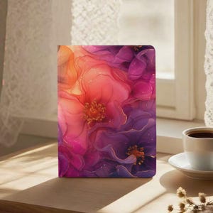 Op de afbeelding: Een bloemrijk notitieboek met een levendig ontwerp. De omslag toont abstracte bloemen in roze, paars en oranje, met gouden accenten. Het notitieboek is rechthoekig en staat op een houten oppervlak.
