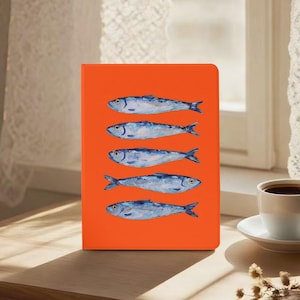 May include: An orange notebook with a watercolour illustration of five blue fish. The fish are arranged vertically on the cover, with details including scales and fins. The notebook is on a wooden surface.