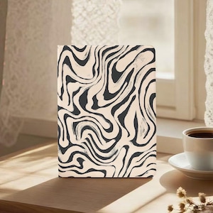 May include: A rectangular notebook with a cream-colored background and a black, abstract, swirled pattern. The notebook is on a wooden surface, with a cup of coffee and a saucer in the background.