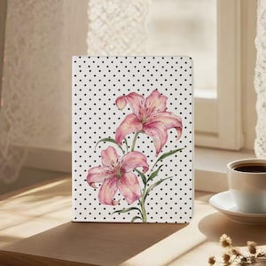 May include: A rectangular notebook with a white cover featuring a black polka dot pattern and a central illustration of two pink lily flowers with green stems and leaves. The notebook is positioned upright on a flat surface.