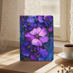 Może przedstawiać: Notatnik z kwiatowym wzorem przedstawiającym duże fioletowe i fioletowe kwiaty. Okładka ma ciemnoniebieskie tło ze złotymi akcentami. Notatnik leży na drewnianej powierzchni w pobliżu okna.