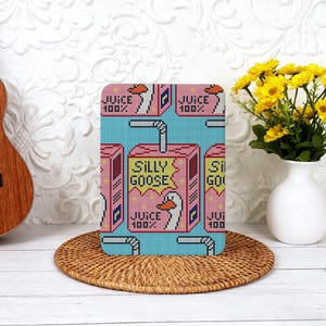 Puede incluir: Un diseño rectangular y pixelado presenta una caja de jugo rosa con las palabras "SILLY GOOSE" y "JUICE 100%" sobre un fondo azul. El diseño está en un objeto rectangular, posiblemente una funda de teléfono o un artículo decorativo.