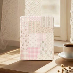 Może przedstawiać: Pastelowo-różowe i białe etui na tablet w stylu patchwork. Etui posiada różnorodne wzory, w tym kwiatowe, w kratkę i w paski. Etui leży na drewnianej powierzchni w pobliżu okna i filiżanki kawy.
