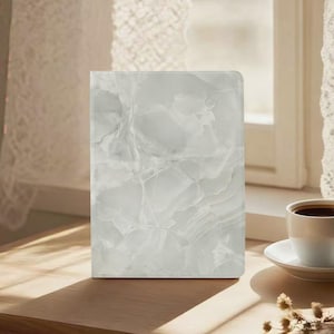 Può includere: Un taccuino rettangolare con una copertina effetto marmo bianco è appoggiato su una superficie di legno. La copertina presenta un design a vortice e venato in bianco e grigio. Una tazza di caffè è nelle vicinanze.