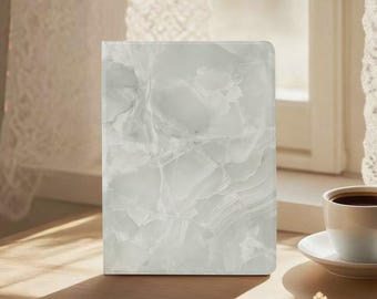 May include: A rectangular, white marble-patterned notebook or journal stands upright on a wooden surface. The cover features a swirling, veined design in shades of white and gray. A cup of coffee sits nearby.