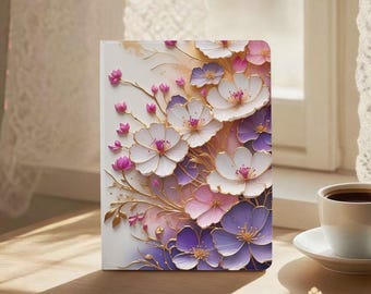 Puede incluir: Un cuaderno blanco con un diseño floral que presenta flores blancas, moradas y rosas con detalles dorados. Las flores están dispuestas en un patrón en capas, creando un efecto 3D. El cuaderno está sobre una superficie de madera.