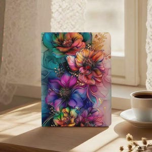 Può includere: Un taccuino con un vivace motivo floreale. La copertina mostra una composizione di fiori nei colori verde acqua, rosa, arancione, viola e giallo. Il taccuino rettangolare è su una superficie di legno, vicino a una finestra.