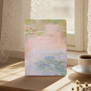 Puede incluir: Un diario de tapa dura con una pintura pastel de nenúfares en un estanque. La obra de arte tiene suaves tonos rosas, verdes y azules. El diario es rectangular y está de pie sobre una superficie de madera, junto a una taza de café.