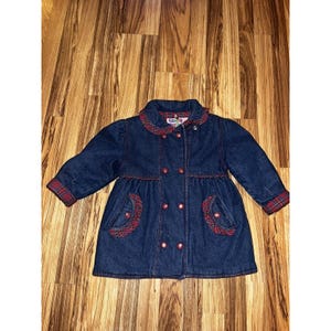 May include: A dark blue denim coat for children with a double-breasted design. The coat features a red and black plaid collar, pocket accents, and cuffs. The coat has a flared skirt and is buttoned up.