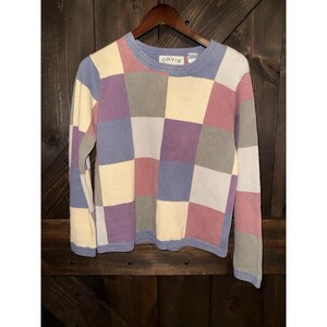 May include: A patchwork sweater in pastel colors, including lavender, pink, cream, and gray squares. The sweater has long sleeves and a crew neck with the brand name "ORVIS" on the collar. The sweater is hanging on a wooden hanger.