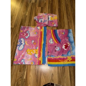 Puede incluir: Juego de ropa de cama rosa de Care Bears con varios personajes de Care Bears, arcoíris y nubes. El juego incluye una sábana bajera, una sábana encimera y una funda de almohada. Las sábanas tienen un fondo rosa con acentos coloridos y gráficos de Care Bears.