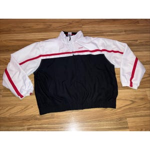 Puede incluir: Chaqueta deportiva Reebok blanca, negra y roja. La chaqueta tiene una sección superior blanca con rayas rojas en el pecho y en las mangas. La sección inferior es negra. La chaqueta tiene cremallera y el logo de Reebok.