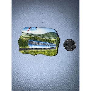 May include: A souvenir magnet featuring a blue train on a green and brown base. The magnet has a white and blue structure on top, with a quarter coin next to it for scale. The train is a light blue color.