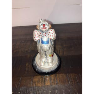 May include: A silver-colored clown figurine with a red nose, curly orange hair, and a small top hat. The clown has a large bow tie with red polka dots and is standing on a black base. The clown's outfit includes a blue vest and suspenders.