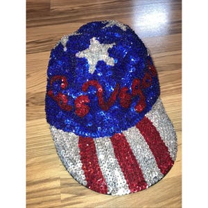May include: A sequined baseball cap with a patriotic design. The cap features a blue crown with a white star and the word "Vegas" in red sequins. The brim has red and silver stripes.