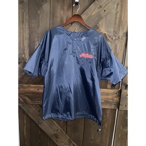 May include: Navy blue short-sleeve baseball jacket with the word "Indians" in red script across the chest. The jacket has a drawstring at the bottom and a button-up front. The jacket is hanging on a wooden hanger.