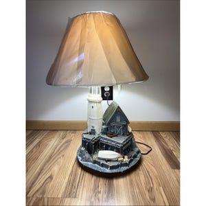May include: A decorative lamp featuring a lighthouse and a small building, with a beige lampshade. The lighthouse is white with a black top, and the building is blue with a green roof. The lamp sits on a wooden base.