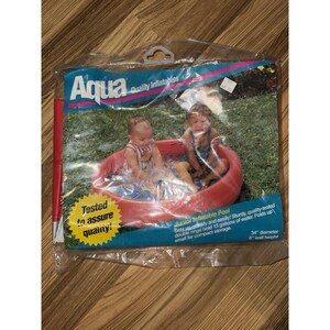 Op de afbeelding: Een verpakte Aqua junior opblaaszwembad. Het rode zwembad, met een diameter van 86 cm en een wandhoogte van 15 cm, heeft twee kinderen die erin spelen. De verpakking bevat de tekst "Getest om kwaliteit te garanderen!"
