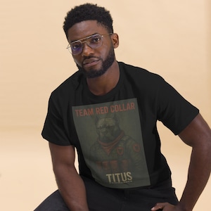 Op de afbeelding: Zwart T-shirt met een afbeelding van een hond met een rode bandana en militair uniform. De tekst "TEAM RED COLLAR" staat boven de afbeelding van de hond en "TITUS" eronder. Het T-shirt wordt door een persoon gedragen.