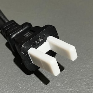 May include: Close-up of a black electrical plug with two white prongs. The plug is connected to a black cord. The image is taken against a gray background.
