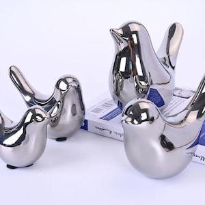 Könnte beinhalten: Eine Sammlung von vier silberfarbenen Keramikvogel-Figuren. Die Vögel sind unterschiedlich groß und auf einer weißen Oberfläche angeordnet, wobei einer auf einem Buch liegt. Die Figuren haben eine glatte, reflektierende Oberfläche und sind mit ausgebreiteten Flügeln gestaltet.