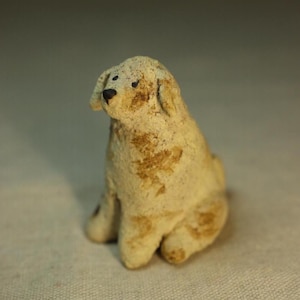 May include: A small, handcrafted ceramic dog figurine in a seated position. The dog is primarily off-white with brown markings and a textured surface. It has a small, dark nose and eyes, and its ears are slightly drooping.
