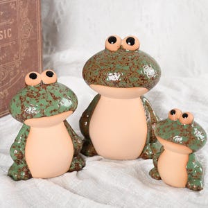 Op de afbeelding: Drie keramische kikkerbeeldjes met een gespikkelde groene glazuur en bruine buiken. De kikkers hebben grote, expressieve ogen en zijn gerangschikt in een familiegroep. Een decoratief boek met de tekst "CLASSIC" staat op de achtergrond.