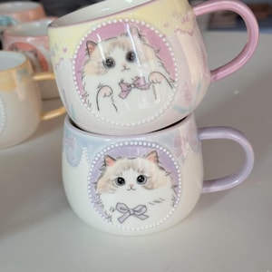 May include: Two stacked ceramic mugs with cat illustrations. The mugs have a white base with a pastel color gradient and a pink handle. Each mug features a cartoon cat with a bow tie in a circular frame.