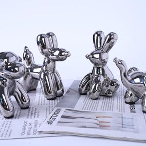 Könnte beinhalten: Vier silberfarbene Ballon-Tier-Skulpturen: ein Hund, ein Pudel, ein Hase und ein Elefant. Die Skulpturen werden auf einer Zeitung präsentiert. Die reflektierenden Oberflächen der Skulpturen erzeugen ein glänzendes Aussehen.