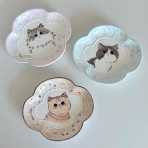 May include: Three decorative ceramic plates with a unique flower-shaped design. Each plate features a hand-painted cat portrait in the centre, surrounded by a decorative border. The plates are in pastel colours: pink, blue, and beige.