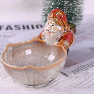 Może przedstawiać: Ceramiczna miseczka w kształcie Świętego Mikołaja z czerwonym kapeluszem i płaszczem oraz białą brodą. Miseczka jest jasnobrązowa z błyszczącym wykończeniem. Słowo "FASHION" jest widoczne w tle.