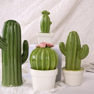 Op de afbeelding: Een verzameling groene keramische cactussculpturen in verschillende vormen en maten. Eén is lang en slank, andere zijn rond of hebben armen. Elke cactus staat in een witte pot, één is bekroond met roze bloemen. Woondecoratie.