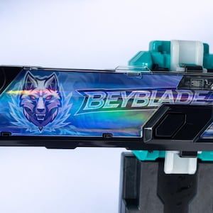 May include: A black and teal Beyblade launcher with a holographic design. The launcher features a wolf graphic and the word "BEYBLADE" in white. The launcher is designed for launching Beyblade tops.