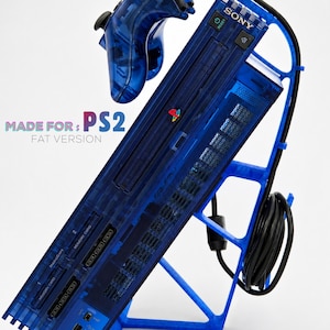 May include: A blue translucent plastic stand holding a Sony PlayStation 2 Fat console and a matching blue translucent controller. The stand features a vertical design with a cable management hook. Text on the image reads: MADE FOR: PS2 FAT VERSION.