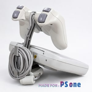 Peut inclure: Un support de manette en plastique gris clair conçu pour une console de jeu, doté d'un bras de support vertical qui maintient une manette et d'un clip circulaire pour organiser le câble gris enroulé. Le texte MADE FOR: PS one est visible.