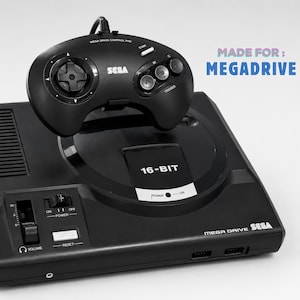 Puede incluir: Una consola de videojuegos Sega Mega Drive negra con un mando con cable a juego. La consola cuenta con una ranura para cartuchos de 16 bits, un control deslizante de volumen y botones de encendido y reinicio. El texto MADE FOR: MEGADRIVE es visible.