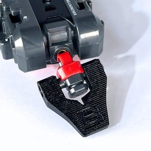 May include: A close-up of a black and gray toy launcher with a red component. The launcher is partially assembled, with a black piece detached. The text "BLADE X STRING LAUNCHER" is visible on the toy.