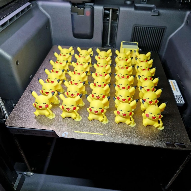 May include: A collection of thirty-five small, yellow, 3D-printed Pikachu figurines arranged in a grid on a dark, textured build plate. Each figure features a smiling expression, red cheeks, and black-tipped ears, all standing in a uniform pose.
