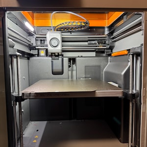May include: A Bambu Lab P2S 3D printer with a dark gray frame, an internal print head, and a square build plate featuring the text 256 x 256 x 256mm. The machine includes a top-mounted digital display screen and internal lighting components.