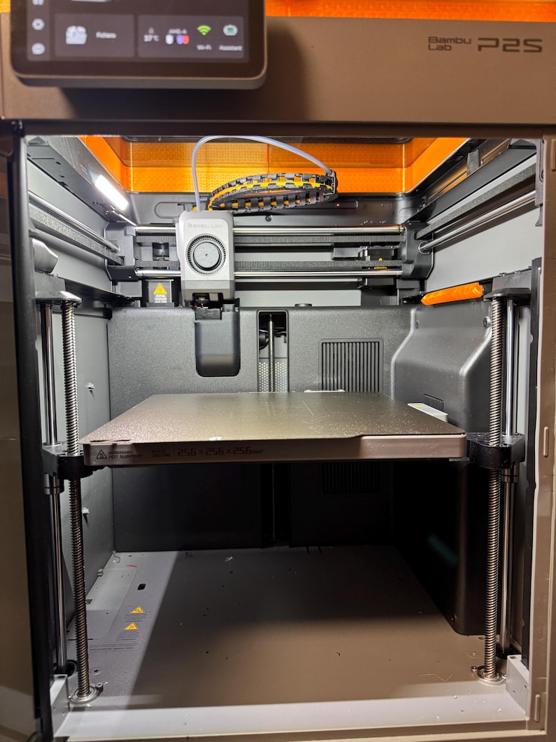May include: A Bambu Lab P2S 3D printer with a dark gray frame, an internal print head, and a square build plate featuring the text 256 x 256 x 256mm. The machine includes a top-mounted digital display screen and internal lighting components.